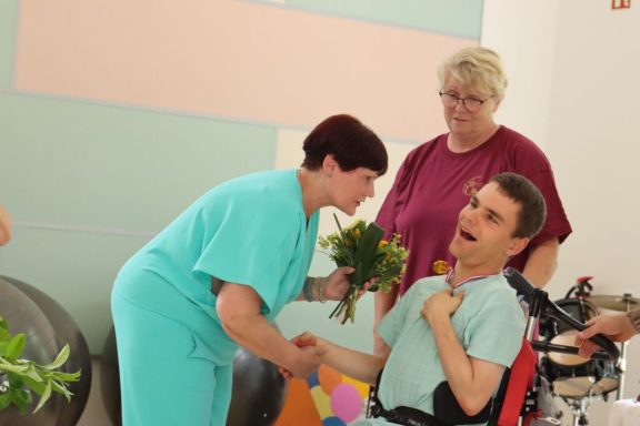 Eine Pflegerin überreicht einem lächelnden Mann im Rollstuhl einen Blumengruß.