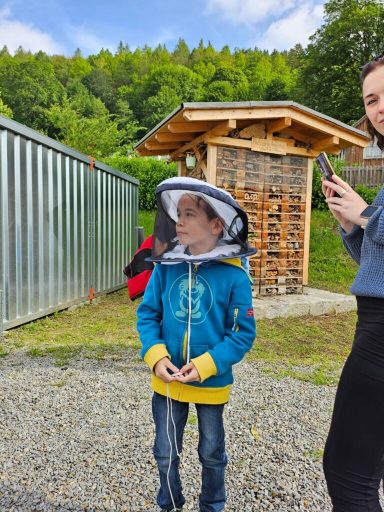 Ein Junge in blauer Jacke mit einem Imkerhut steht im Freien, während eine Frau am Handy steht.