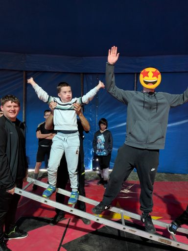 Zwei Personen halten einen Jungen auf einer Wippe in einem Zirkuszelt.