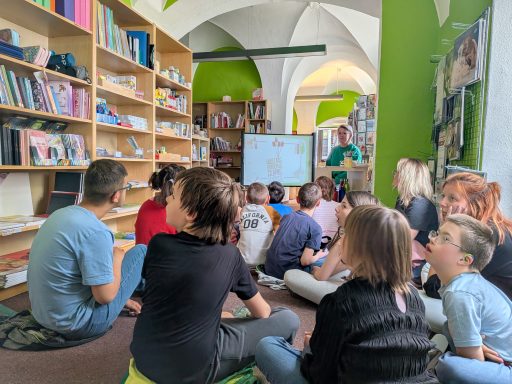 Gruppe von Kindern sitzt in einem Raum und schaut auf eine Präsentation an einer Leinwand.