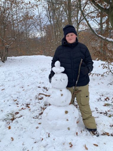 Person im Schnee steht neben einem Schneemann, umgeben von Bäumen und Schnee.