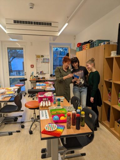 Drei Frauen stehen in einem hellen Raum, umgeben von Bastelmaterialien und Tischen.