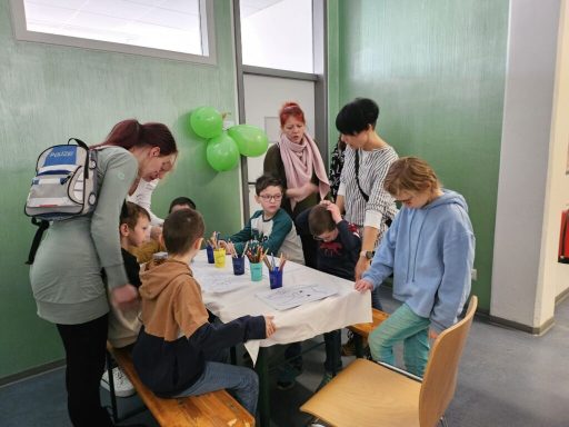 Kinder sitzen an einem Tisch und malen, umgeben von Erwachsenen und grünen Ballons.