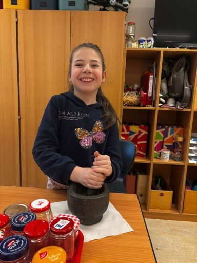 Ein Mädchen steht lächelnd mit einem Mörser und stößt Zutaten im Hintergrund.