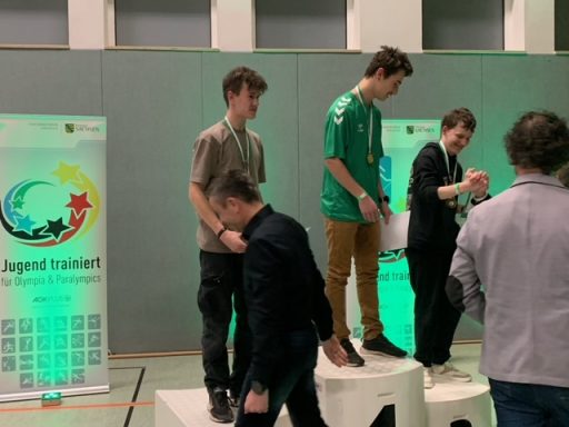 Zwei Jugendliche stehen auf einem Podest, während eine Person daneben geht. Hintergrund mit Banner.