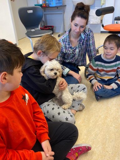 Vier Kinder sitzen am Boden, während eine Frau mit einem kleinen Hund sitzt und lächelt.
