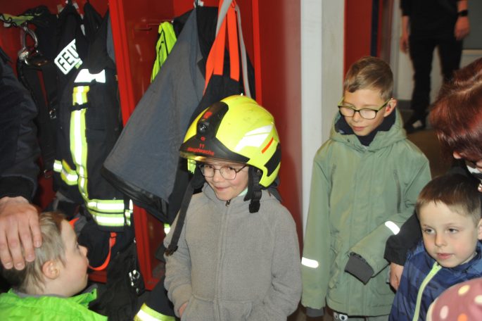 Kinder stehen in einer Feuerwehrstation, einer trägt einen Feuerwehrhelm.