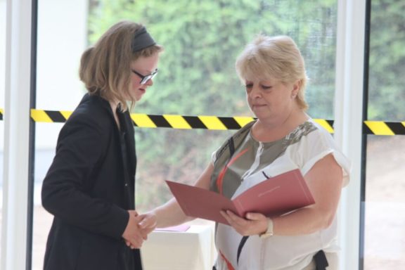 Zwei Frauen schütteln sich die Hände und halten Dokumente in einem hellen Raum.