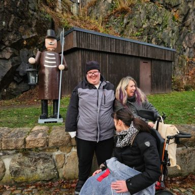 Drei Personen posieren vor einem Holzfigur in einem herbstlichen Landschaft.