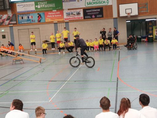 Ein BMX-Fahrer zeigt Tricks vor Publikum in gelben T-Shirts in einer Sporthalle.