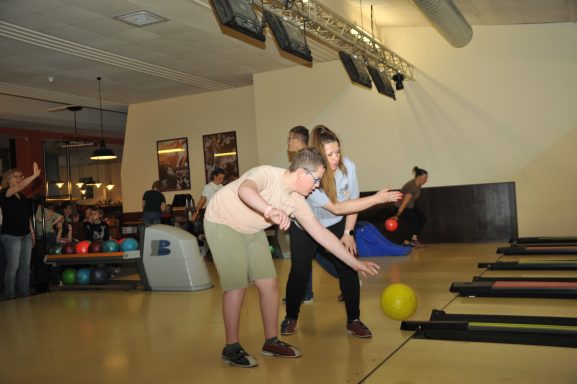 Drei Personen spielen Bowling in einer Halle, während eine Kugel gerollt wird.