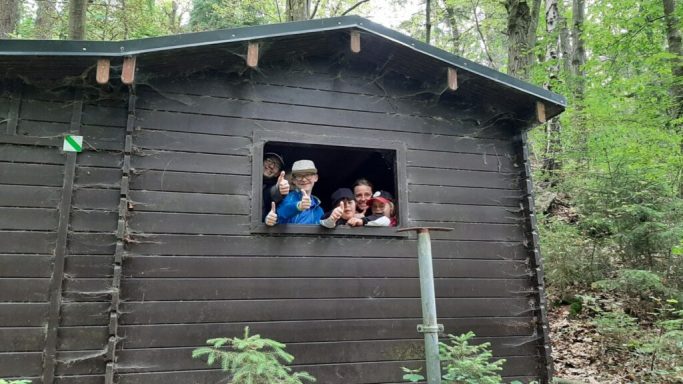 Zwei Personen winken aus einem Fenster einer Holzhütte im Wald.