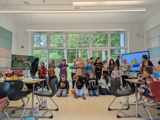Kinder stehen in einem hellen Klassenraum, umgeben von Tischen und Fenstern.