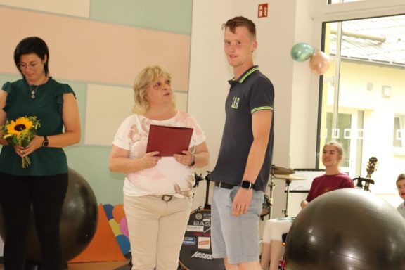 Zwei Personen stehen im Vordergrund, eine hält ein Dokument, im Hintergrund sind weitere Menschen.