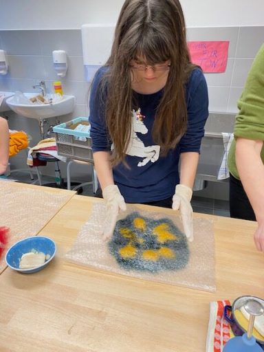 Eine Person mit langen Haaren bereitet mit Handschuhen Lebensmittel auf einem Tisch vor.