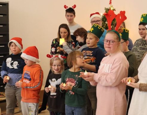 Grupp von Kindern und Erwachsenen in festlichen Hüten, lächeln und halten Kerzen.