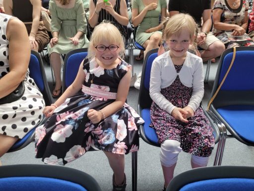 Zwei Mädchen in festlicher Kleidung sitzen auf blauen Stühlen in einem Saal.