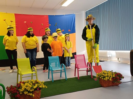 Gruppenvorführung mit Kinder in bunten Kostümen und Stühlen auf einer Bühne.