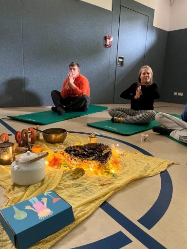 Zwei Personen in Meditationshaltung auf Matten, umgeben von Dekoration und einer Schale.