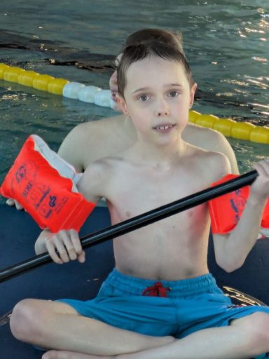 Ein Junge mit Schwimmflügeln sitzt auf einer Matte am Poolrand.