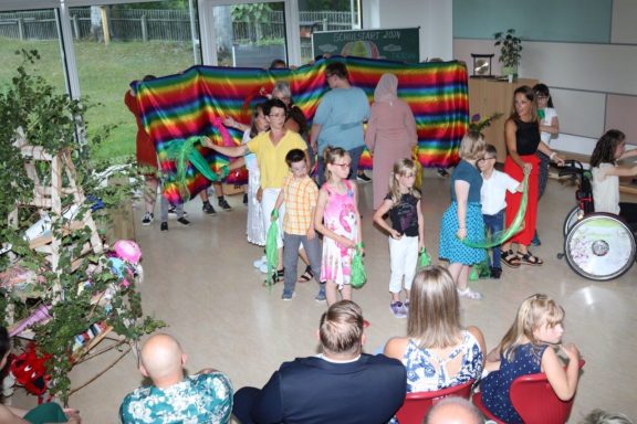 Kinder stehen in bunten Outfits auf einer Bühne vor einem farbenfrohen Hintergrund.