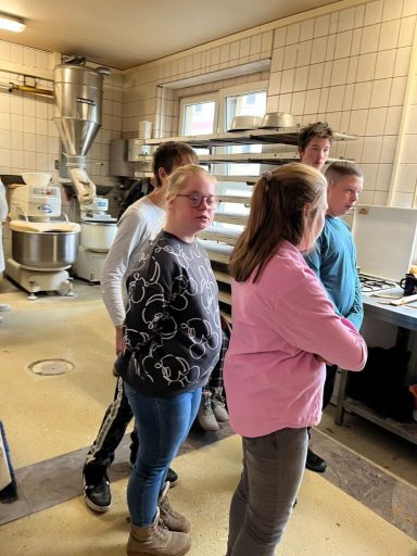Fünf Personen stehen in einer Bäckerei, einige schauen zur Theke, andere miteinander.