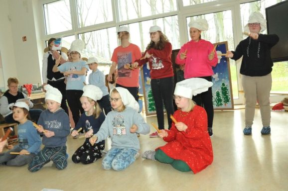 Kinder und Erwachsene in Küchenmützen führen eine Aufführung in einem hellen Raum auf.