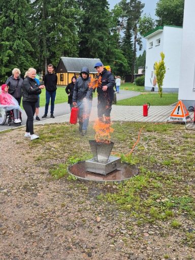 Person in Feuerwehruniform demonstriert das Löschen eines Feuers, umgeben von Zuschauern.