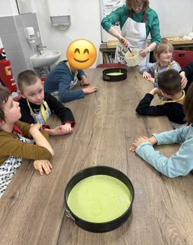 Gruppe von Kindern sitzt um einen Tisch, beobachtet das Backen mit grüner Kuchenmasse.