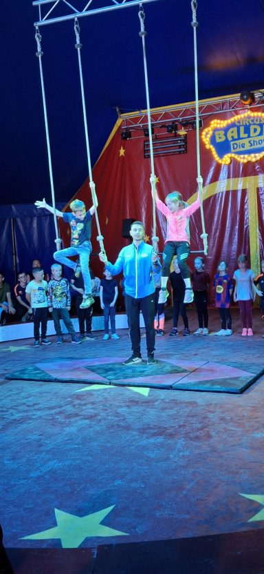 Zwei Kinder schwingen an Seilen, ein Erwachsener steht in der Mitte, Publikum im Hintergrund.