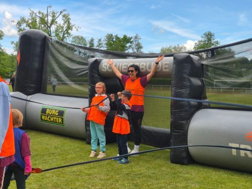 Kinder in orange Westen stehen vor einem großen Spielgerät im Freien.