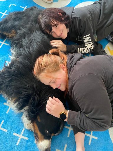 Zwei Frauen liegen auf einem Teppich und kuscheln mit einem großen Hund.