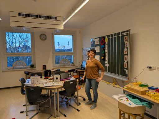 Eine Person steht in einem Klassenraum mit Tischen und Lehrmaterialien.