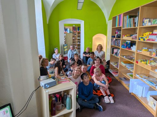Gruppe von Kindern und Erwachsenen in einem bunten Raum mit Bücherregalen und grünen Wänden.