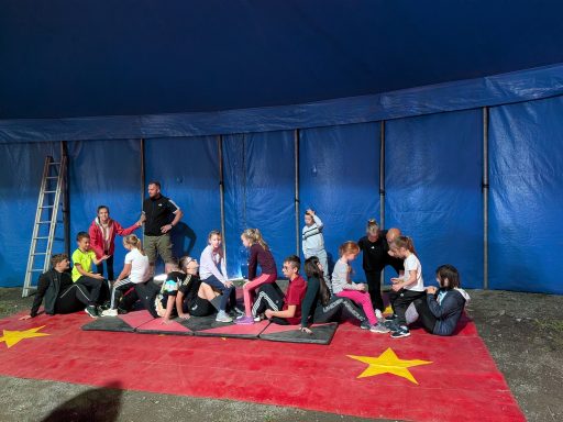 Es sitzen mehrere Personen auf einem roten Teppich in einem Zirkuszelt.