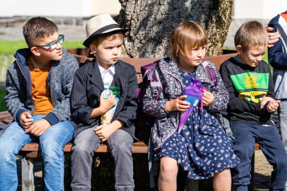 Vier Kinder sitzen auf einer Bank, tragen unterschiedliche Kleidung und schauen nach vorne.