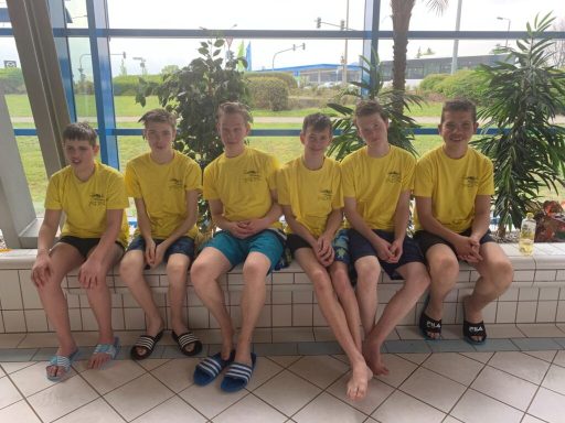 Sechs Jugendliche in gelben T-Shirts sitzen auf einer Bank in einer Schwimmhalle.