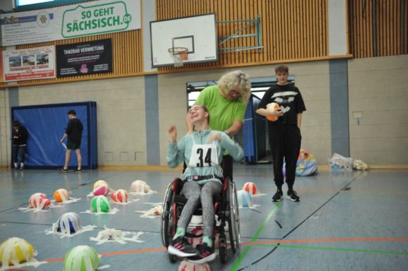 Eine Person im Rollstuhl lacht, während eine andere sie anfeuert. Bunte Bälle liegen um sie herum.