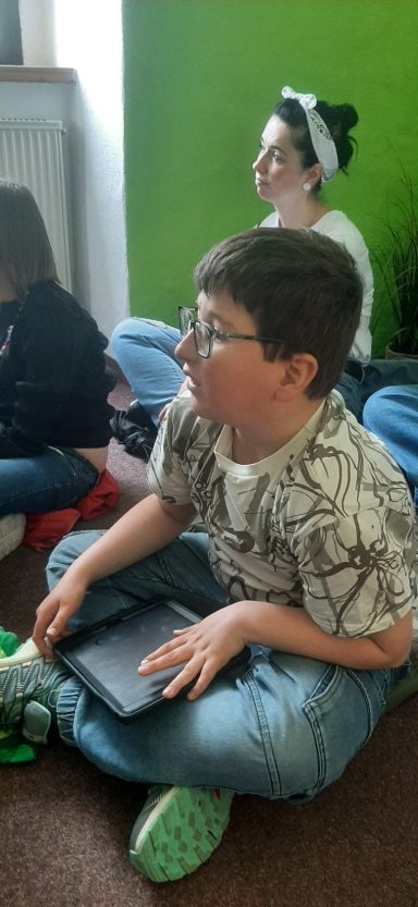 Ein Junge mit Brille sitzt auf dem Boden und hält ein Tablet, während andere im Hintergrund sitzen.