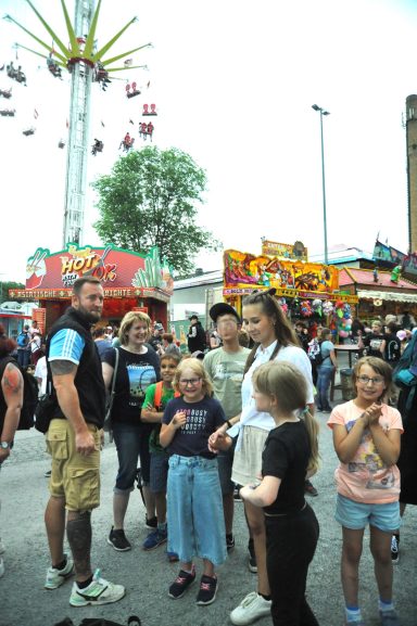 Gruppierung von Menschen auf einem Jahrmarkt mit Fahrgeschäften im Hintergrund.