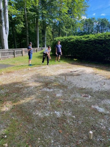 Drei Kinder spielen im Freien auf einem kiesigen Boden vor bunten grünen Büschen.
