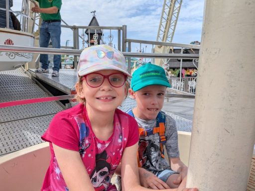 Zwei lächelnde Kinder auf einem Fahrgeschäft, umgeben von Fahrgeschäften im Hintergrund.