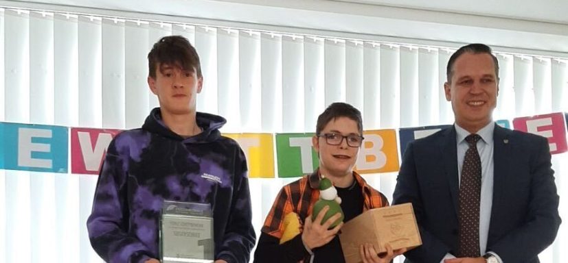 Zwei junge Preisträger halten Urkunden, umgeben von einem Erwachsenen und festlichem Dekor.