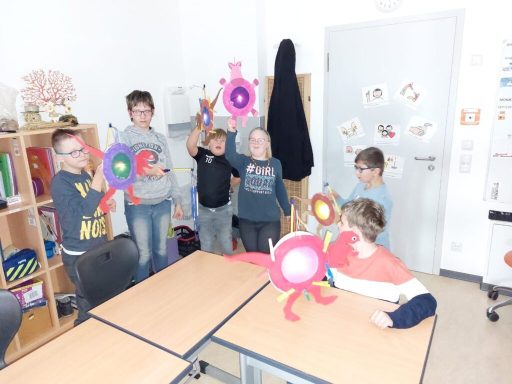 Kinder stehen in einem Klassenzimmer und halten bunte Bastelarbeiten hoch.