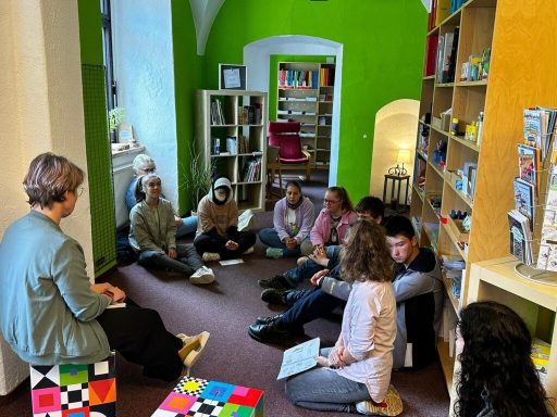 Gruppendiskussion in einem hellen Raum mit Regalen voller Bücher und Sitzkissen.