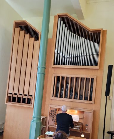 Ein Musiker spielt an einer Holzorgel mit großen Pfeifen in einem lichtdurchfluteten Raum.