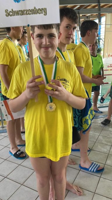 Junger Schwimmer in gelbem T-Shirt mit Medaille, hält ein Schild mit "Schwimmjunge".