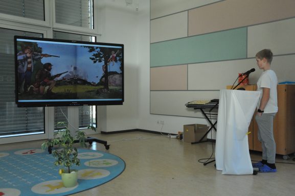 Ein Junge steht an einem Rednerpult vor einem großen Bildschirm mit einer Präsentation.