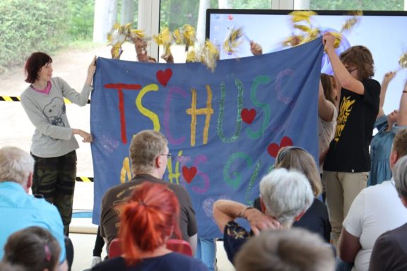 Gruppendarstellung mit buntem Banner, auf dem "TSCHÜS" steht, umringt von Zuschauern.