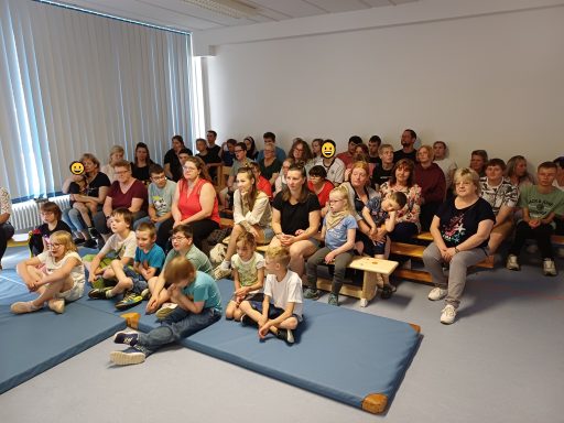Eine Gruppe von Menschen, darunter viele Kinder, sitzt in einem Raum und schaut nach vorne.